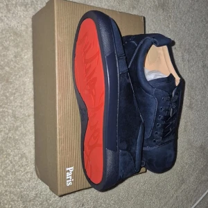 Mörkblå sneakers från Christian Louboutin - Snygga mörkblå sneakers från Christian Louboutin i mocka med klassisk röd sula. Skorna har rund tå, platt sula och snörning. Insidan är i beige läder och de kommer med originalkartong och dustbag. Perfekta för dig som vill sticka ut med exklusiva detaljer.