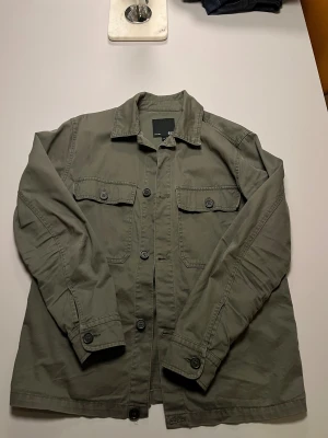 Grön overshirt med knappar - Snygg militärgrön overshirt från 153 med klassisk krage och stora bröstfickor med knappar. Jackan har lång ärm och stängs med knappar framtill. Perfekt att slänga över en hoodie eller t-shirt för en chill look.