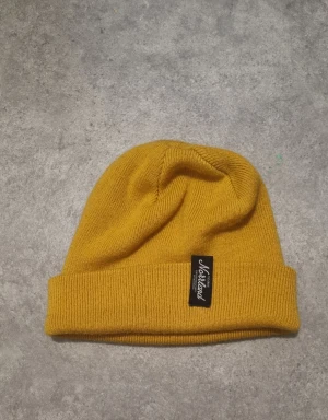 Gul stickad mössa från Norrländ - Snygg gul mössa från Norrländ med uppvikt kant och svart tygmärke framtill. Stickad i mjukt material som håller dig varm och ger en chill vibe. Perfekt för dig som vill ha en färgklick till din outfit.