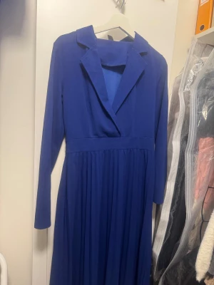 Kornblå långklänning från SHEIN - Kornblå långklänning från SHEIN med lång ärm, markerad midja och plisserad kjol. Klänningen har en snygg krage och omlottliknande överdel. Perfekt för dig som vill sticka ut med en elegant och stilren look.