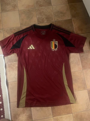Belgien fotbollströja Adidas  - Säljer en officiell Belgien fotbollströja från Adidas i vinrött med svarta och guldiga detaljer. Tröjan har korta ärmar, mönstrad framsida och broderat landslagsemblem på bröstet. Materialet är lätt och andas, perfekt för match, träning eller att bara ha på sig den.