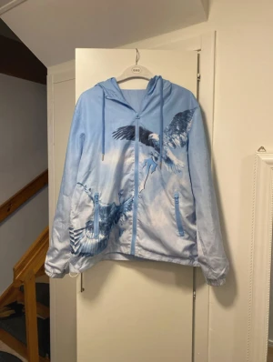 Trapstar blå vindjacka med örnmotiv - Säljer en blå vindjacka från Trapstar med snyggt örnmotiv och tryckt logga på ryggen. Jackan har huva med snörning, dragkedja framtill och två fickor med dragkedja. Tillverkad i 100% polyester, perfekt för dig som vill sticka ut med streetwear.