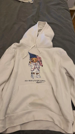 Vit hoodie Polo Bear Ralph Lauren - Vit hoodie från Polo Ralph Lauren med ikoniska Polo Bear-trycket på bröstet där björnen håller en amerikansk flagga. Tröjan har huva med snörning och stor magficka. Mjuk bomullskvalitet och klassisk passform, perfekt för chill dagar.skriv gärna om du har några frågor.priset kan diskuteras.