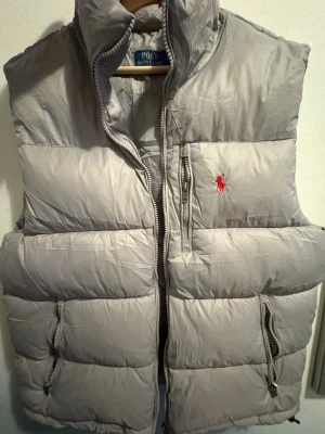 Grå väst från Polo Ralph Lauren - Snygg grå väst från Polo Ralph Lauren med hög krage och klassisk röd logga broderad på bröstet. Västen har flera dragkedjeförsedda fickor och quiltad design som ger en puffig look. Perfekt för lager-på-lager-stil under kyliga dagar.