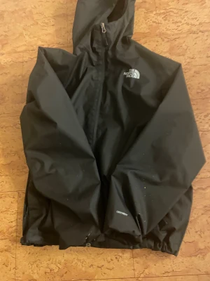 Svart vindjacka från The North Face - Svart vindjacka från The North Face med huva och dragkedja framtill. Jackan har meshfoder på insidan och logga på bröstet. Perfekt för blåsiga dagar och har en clean, sportig look. Modellen är klassisk och passar dig som gillar streetwear.