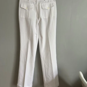 Vita byxor från Wear It - Snygga vita byxor från Wear It i rak modell med två fickor med lock och knapp framtill. Byxorna har en klassisk passform och är tillverkade i ett mjukt bomullsmaterial. Perfekta för dig som gillar stilrena och fräscha looks. De är något genomskinliga men inte mycket, har även sytt de mer lågmidjade