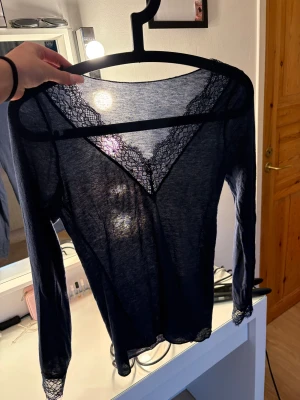 Mörkblå spetsblus från Intimissimi - Säljer en tunn och mörkblå blus från Intimissimi med v-ringad spetsdetalj framtill och vid ärmslut. Blusen är långärmad och har en lätt transparent look som ger en cool vibe. Perfekt för dig som gillar stilrena och feminina detaljer.