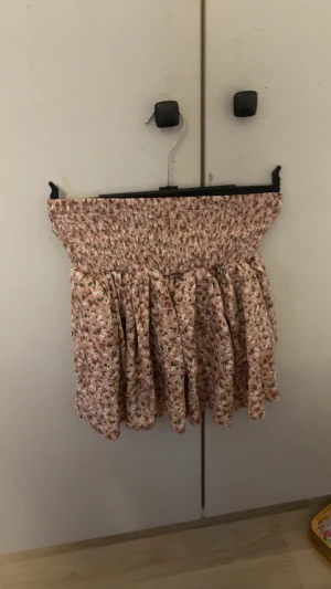 Blommig kortkjol med volang - Supersöt kortkjol i ljusrosa med små blommor i grönt och vitt. Kjolen har smock i midjan för en bekväm passform och volangig nederdel som ger en luftig känsla. Perfekt för varma dagar och en romantisk look.