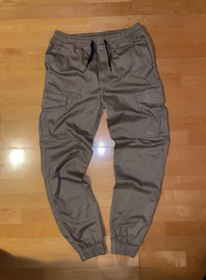 Bruna/ Beige cargopants från Vailent, M - Snygga bruna/beige cargopants från Vailent med elastisk midja och svarta snören. Köpta på carlings för ett tag sedan men aldrig använda. Byxorna har flera praktiska fickor med lock och knappar på sidorna och bak, samt resår vid bensluten för en avslappnad look. Jag skulle säga att de är mer beige i verkligheten än på bilden. Perfekta för en chill och streetinspirerad stil. Har för mig att de kostade ca 599-699 nya. Skriv vid minsta fråga!