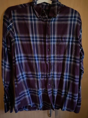 Rutig lila och blå skjorta - Snygg rutig skjorta i lila och blå toner med klassisk krage och knappar framtill. Skjortan har lång ärm och en bröstficka, perfekt för dig som gillar avslappnad stil med lite edge. Materialet känns mjukt och bekvämt mot huden.