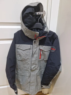 Grå och svart vinterjacka Burton L - Säljer en grå och svart vinterjacka från Burton i barn storlek L 150-164, till åldrarna 14-16. Jackan har huva, flera fickor med lock och dragkedja i rött, samt ett snyggt Burton-märke på ärmen. Insidan är fodrad med mjuk fleece och jackan har justerbara muddar. Aldrig används.