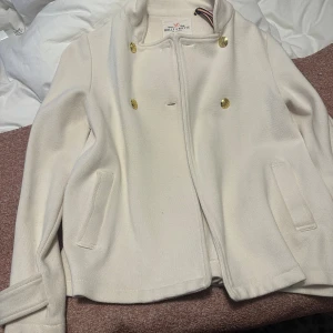 Vit kavajjacka från Holly & White - Snygg vit kavajjacka från Holly & White med guldknappar framtill och på ärmsluten. Jackan har en klassisk rak passform, två fickor och en slits bak. Materialet är mjukt och känns lyxigt, perfekt för dig som gillar stilrena detaljer.