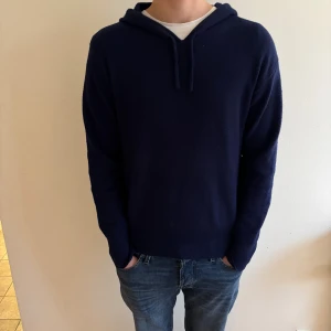 Mörkblå hoodie i kashmir - Snygg mörkblå hoodie i mjuk kashmir med huva och långa ärmar. Tröjan har en klassisk passform och ribbade muddar vid ärmslut och nederkant. Perfekt för dig som gillar stilrena och bekväma plagg med lite lyxig känsla.