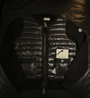 Svart quiltad jacka från Moncler - Snygg svart quiltad jacka från Moncler med blank front och stickade ärmar. Jackan har dragkedja, rund halsringning och två fickor framtill. Klassisk Moncler-logga på ärmen och coola detaljer som ger en exklusiv vibe.