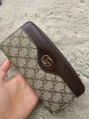 plånbok med GG-mönster - Snygg plånbok med klassiskt GG-mönster i beige och brunt. Plånboken har bruna skinnpaneler, guldfärgat GG-emblem och dragkedja runt om. Insidan har flera fack för kort och mynt. Perfekt för dig som gillar lyxiga detaljer och stilren design.