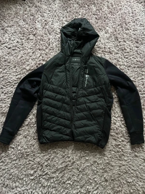 Svart puffer ”cardigan” från Sail Racing - Svart cardigan från Sail Racing med luva och snyggt quiltad framsida. Jackan har dragkedja, långärmade släta ärmar och logga tryckt på bröstet. Perfekt för kyliga dagar. Sportig och clean look. Tveka inte att höra av er vid funderingar eller för fler bilder ☺️bvsa