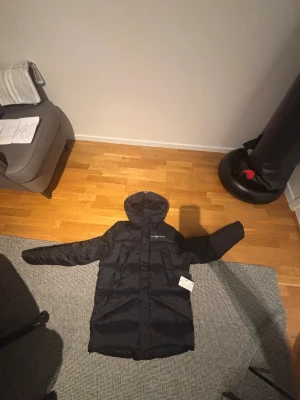 Svart lång dunjacka Sail Racing - Svart lång dunjacka från Sail Racing med huva och diskret logga på bröstet. Jackan har en clean look, dragkedja framtill och två sidofickor. Perfekt för kalla dagar med sin fluffiga och värmande design. Passar dig som gillar stilren och funktionell outerwear. Finns kvitto ny pris cirka 5000