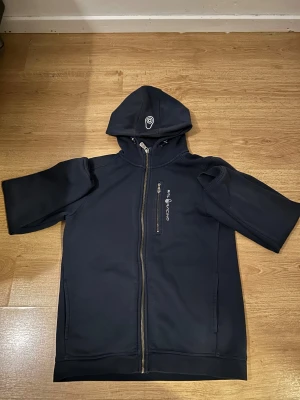 Marinblå hoodie från Sail Racing - Snygg marinblå hoodie från Sail Racing med korta ärmar, hel dragkedja och huva med logga. Tröjan har en bröstficka med dragkedja och Sail Racing-tryck. Materialet är mjukt och perfekt för chill dagar. Passformen är normal och stilen är sportig.