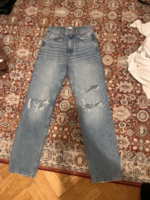  ljusblå jeans med slitningar - Säljer ett par klassiska jeans i ljusblå tvätt med raka ben och hög midja. Jeansen har snygga slitningar och hål på båda benen framtill för en cool, avslappnad vibe. Tillverkade i robust bomullsdenim med fem fickor och knappgylf.