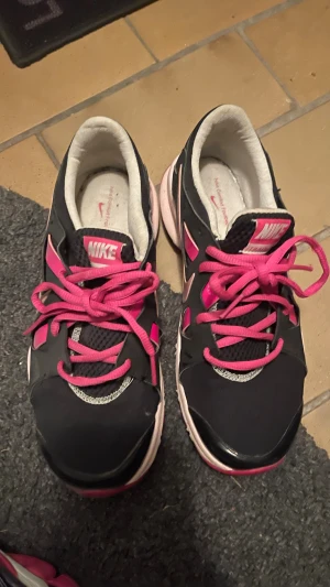 Svarta och rosa Nike sneakers strl 38 - Nike sneakers i svart med rosa detaljer och snörning. Skorna har en rund tå och platt sula, med meshpartier för extra ventilation. Insidan är ljus och mjuk för komfort. Perfekt för dig som gillar sportig stil och färgstarka detaljer.