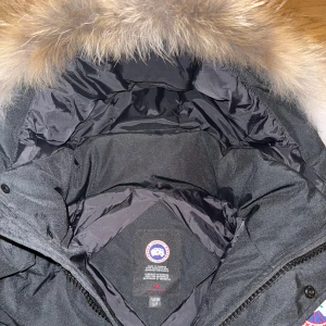 Canada Goose wyndham jacket  - Säljer en svart Canada Goose dunjacka med stor fluffig päls på huvan och klassisk röd logga på ärmen. Jackan har dragkedja och knappar framtill, samt snörning vid huvan. Perfekt för kalla vinterdagar och riktigt snygg streetstil.