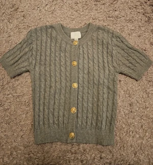 Grå kabelstickad kofta från H&M - Grå kabelstickad kofta från H&M med korta ärmar och snygga guldknappar framtill. Klassisk rund halsringning och ribbade muddar. Mycket bra skick, använd endast en gång. 