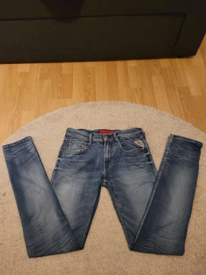Replay Anbass blå jeans slim fit - Snygga Replay Anbass jeans i blå tvätt med få slitningar och klassiska fem fickor. Slim fit-modell med smal passform och normal midja. Jeansen har Replay-logga på bakfickan och röd etikett vid framfickan. Perfekt för dig som gillar stilrena och moderna jeans.