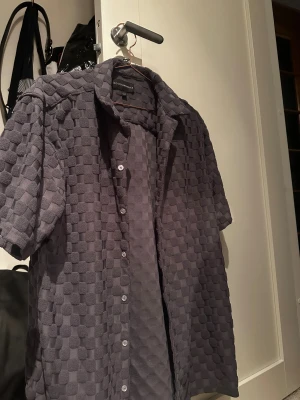 Grå kortärmad skjorta No Boundaries - Säljer en grå skjorta från No Boundaries med korta ärmar och coolt rutigt strukturmönster i froté tyg. Skjortan har knappar framtill och en oversized vibe. Perfekt för dig som gillar unika texturer och vill sticka ut lite extra.