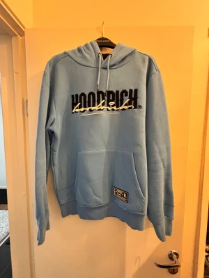 Ljusblå hoodie från Hoodrich - Snygg ljusblå hoodie från Hoodrich med stor logga broderad på bröstet och tryck på ryggen. Tröjan har huva med snörning, stor magficka och ribbade muddar. 