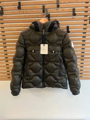 Mörkgrön Moncler dunjacka  - Snygg mörkgrön dunjacka från Moncler med huva och cool quiltad design. Jackan har två framfickor med lock, kraftig dragkedja med Moncler-logga och detaljer i svart. Perfekt för dig som vill ha en varm och stilren jacka med exklusiv känsla. Fitter str s men er 14a jeg er 175 og den passer mig godt