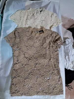 2 spetsblusar från Zara, beige & vit - Två kortärmade spetsblusar från Zara i storlek S. En i beige och en i vit, båda med hög hals och vackert blommigt spetsmönster. Blusarna har knapp i nacken och genombruten design som ger en romantisk vibe. Xs/s kan vara liten i storlek