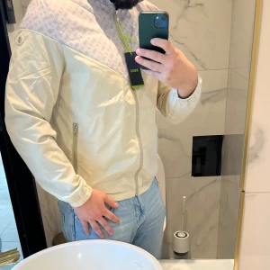 Beige vindjacka från BOSS - Snygg beige vindjacka från BOSS med ljusgrått mönstrat parti över axlar och bröst. Jackan har dragkedja framtill, två sidofickor med dragkedja och elastiska muddar. BOSS-logga på ärmen och coolt mönster upptill. Perfekt för dig som gillar stilren design.