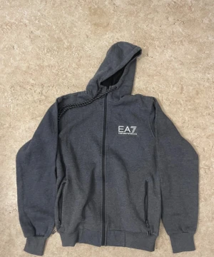 Mörkgrå EA7 hoodie med dragkedja - Snygg mörkgrå hoodie från EA7 Emporio Armani med hel dragkedja framtill, två fickor med dragkedja och justerbar huva med snören. Tröjan har ribbade muddar och en stilren logga på bröstet. Perfekt för en chill och sportig look.