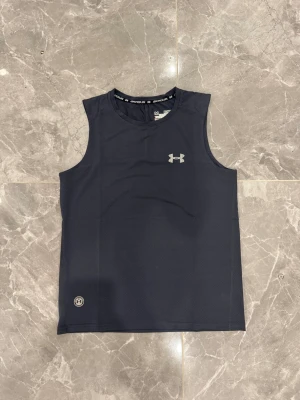 Mörkblå träningstopp från Under Armour - Säljer en mörkblå ärmlös träningstopp från Under Armour med diskret meshstruktur och logga i silver på bröstet. Toppen är tillverkad i syntetmaterial med kompressionspassning och har rund hals. Perfekt för gymmet eller löpning!