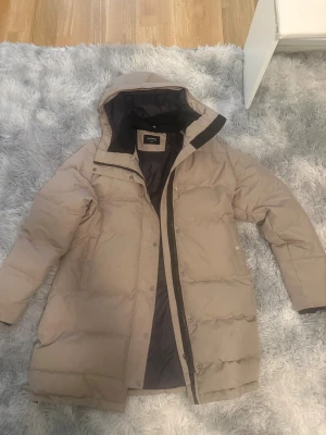 Beige medellång puffervinterjacka från Everest - Säljer en beige puffervinterjacka från Everest med huva och långa ärmar. Jackan är medellång, har dragkedja och tryckknappar framtill samt två fickor. Insidan är svart och materialet är syntet. Perfekt för kalla dagar när du vill hålla dig varm och stilsäker.