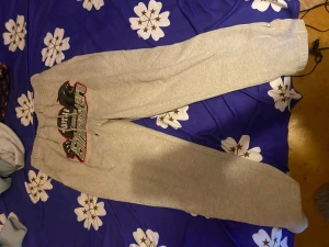 Grå mjukisbyxor med tryck och dragkedja - Snygga grå mjukisbyxor med stort tryck framtill och coolt svart/rött motiv. Byxorna har snörning i midjan och dragkedja vid bensluten för extra stilpoäng. Perfekta för chill eller träning, mjukt material och relaxed passform.