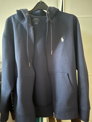Marinblå hoodie från Polo Ralph Lauren - Snygg marinblå hoodie från Polo Ralph Lauren med klassisk vit logga på bröstet. Tröjan har huva med snörning, dragkedja framtill och två rymliga fickor. Perfekt för chill dagar och enkel att matcha med jeans eller joggers.