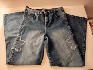 Blå jeans med stjärndetaljer - Säljer ett par blå jeans med breda ben och coola slitna stjärnor på både fram- och baksidan av benen. Jeansen har klassisk femficksdesign och är tillverkade i jeansmaterial. Perfekt för dig som vill sticka ut med en unik look. Köpte det utomlands så den har inget märke.