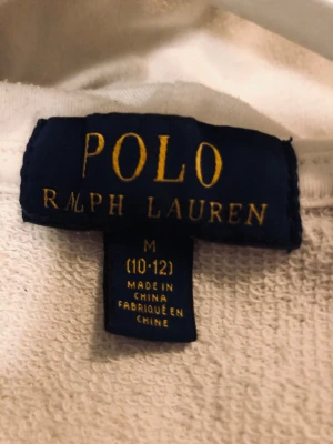Vit hoodie från Polo Ralph Lauren - Vit hoodie från Polo Ralph Lauren med stor röd POLO-text framtill. Tröjan har dragkedja, huva och två fickor. Tillverkad i mjuk bomull, perfekt för chill dagar. Klassisk och enkel stil som funkar året runt.