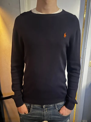 Mörkblå stickad tröja från Ralph Lauren - Snygg mörkblå stickad tröja från Ralph Lauren med klassisk rund hals och orange broderad logga på bröstet. Strlk M, slimfit, och modellen på bilden är 193 och väger 82kg. Köpt för 1200, pris kan diskuteras