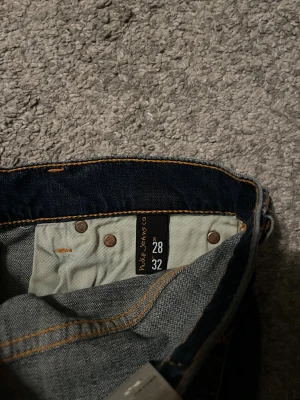 Mörkblå jeans från Nudie Jeans Co W28 L32 - Snygga mörkblå jeans från Nudie Jeans Co i klassisk femficksmodell. Jeansen har en rak passform med subtila slitningar och kontrastsömmar. Materialet är robust denim i bomull och bakfickan har Nudie Jeans signaturbroderi.