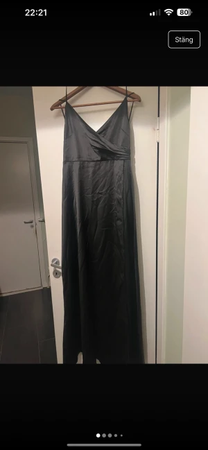 Svart långklänning med omlott - Svart långklänning med smala axelband och omlott framtill. Klänningen har en elegant v-ringning och är tillverkad i ett glansigt, mjukt material (satin) som ger en lyxig känsla. Öppen lite där bak med snörning Perfekt för dig som vill ha en stilren och klassisk look. Kanske din nya nyårsklänning? Den är oanvänd. 