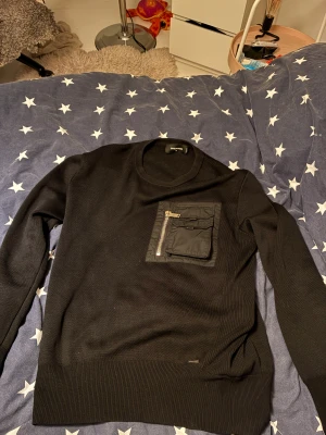 Svart Dsquared2 tröja  - Svart långärmad tröja från Dsquared2 i storlek XL men sitter som L. Tröjan har en unik ficka med dragkedja och lock på bröstet, vilket ger en cool och modern vibe. Materialet är stickat och känns mjukt mot huden. Perfekt för dig som gillar stilrena detaljer.