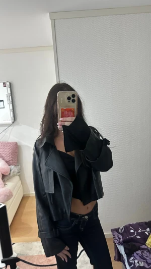 Svart croppad skinnjacka med bred krage - Säljer en svart croppad skinnjacka med bred krage och coola detaljer vid ärmsluten. Jackan har en loose passform och ger en edgy vibe till din outfit. Perfekt för dig som gillar streetstyle och vill sticka ut med en snygg jacka i syntetiskt skinn.