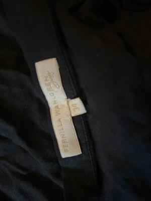 Svart omlottkjol Pernilla Wahlgren - Säljer en svart lång omlottkjol från Pernilla Wahlgren. Kjolen har volangkant nertill och knytband i midjan för en snygg och justerbar passform. Perfekt för dig som gillar en enkel men ändå trendig look.