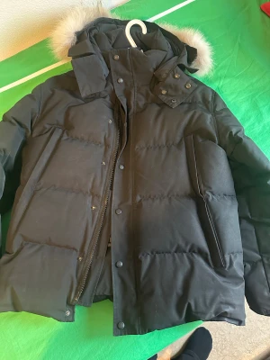 Svart Canada Goose dunjacka med päls - Säljer en svart dunjacka från Canada Goose med fluffig päls på huvan. Jackan har dragkedja och knappar framtill, samt två stora fickor. Märket syns på ärmen och jackan har en schysst, puffig look som är perfekt när det är kallt ute. Passar L också 