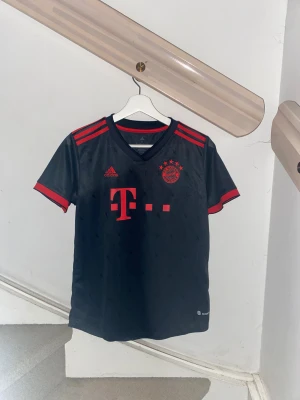 FC Bayern München Adidas t-shirt - Snygg svart FC Bayern München fotbollströja från Adidas med röda detaljer, klubbmärke och sponsortryck fram. Müller #25 på ryggen. Tillverkad i lätt och ventilerande material, perfekt för match eller träning. Klassisk V-ringning och korta ärmar.