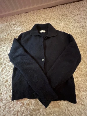 Svart kofta från American Vintage - Mysig svart kofta från American Vintage med klassisk krage och knäppning framtill. Koftan är stickad och har långa ärmar samt ribbade muddar. Perfekt för lager-på-lager och chill vibes.