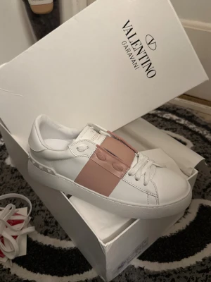 Valentino Garavani Open sneakers vit/rosa - Helt nya & oanvända, snygga Valentino Garavani Open sneakers i vitt skinn med bred ljusrosa läderdetalj över snörningen. Platt sula med mönster och ikoniska nitar baktill. Rund tå och klassisk siluett, perfekt för dig som gillar exklusiv streetstyle.