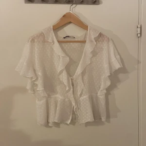 Vit prickig blus med volanger från Zara - Säljer en söt vit blus från Zara i storlek M. Blusen har ett prickigt mönster, volanger längs kanterna och korta ärmar. Den är lätt och luftig, passar super över ett vitt linne eller annat basplagg🩷🩷